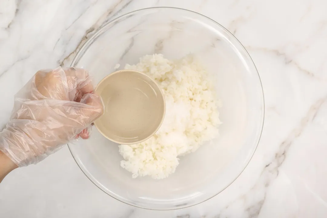 a hand holds a small bowl of water on top of a glass bowl of cooked rice