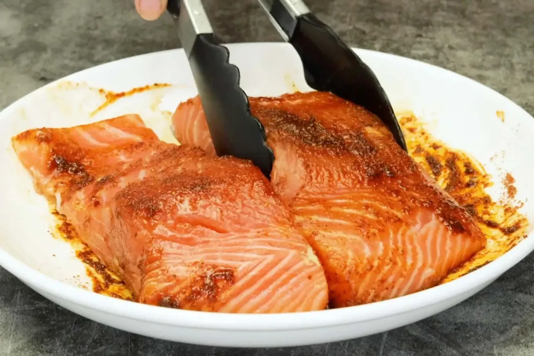 A pair of tongs is touching one out of the two salmon fillets topped with seasonings on a white plate.