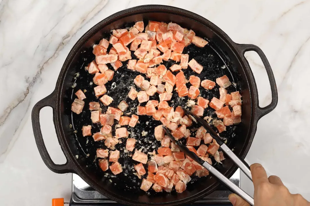 Salmon chunks stirred in a cast iron skillet with a pair of tongs
