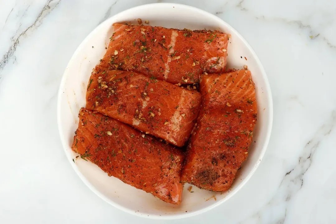 A round white plate containing four uncooked salmon fillets covered in spices and herbs