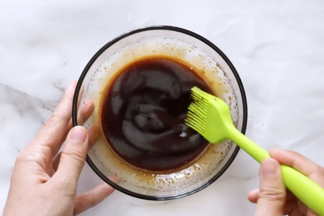 Two hands mixing a glossy brown sauce in a small glass bowl with a neon green butter brush
