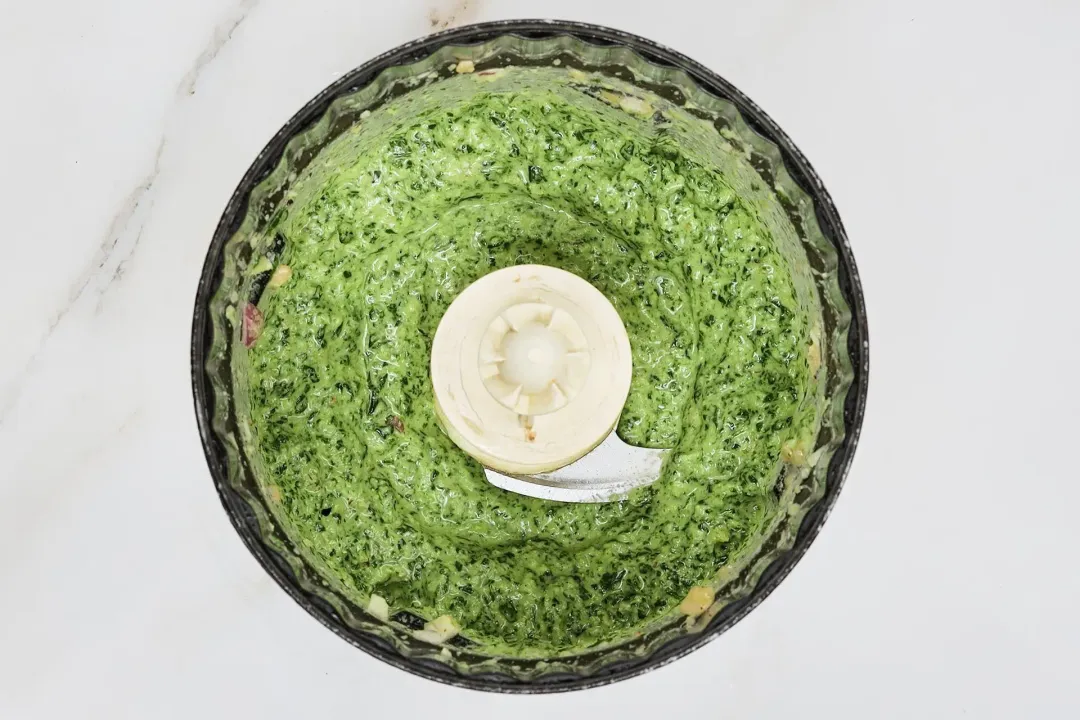 A picture of blanched spinach, basil, garlic cloves, shallot and seasonings being blended in a food processor