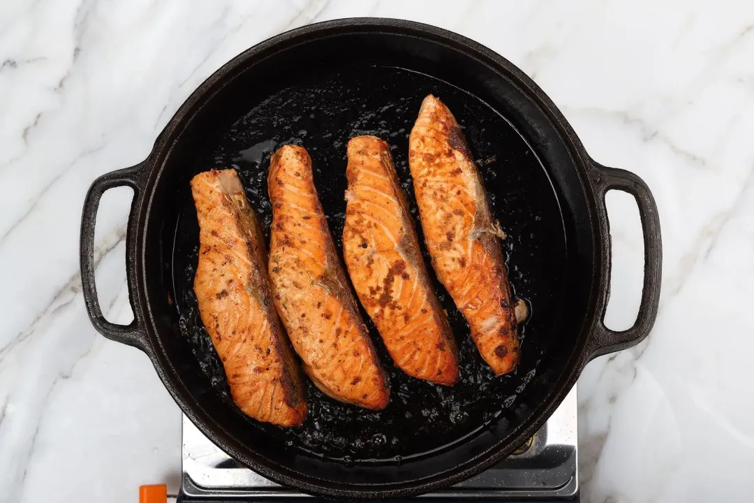 A skillet cooking four salmon fillets. Their exterior is bright orange and have little brown spots.