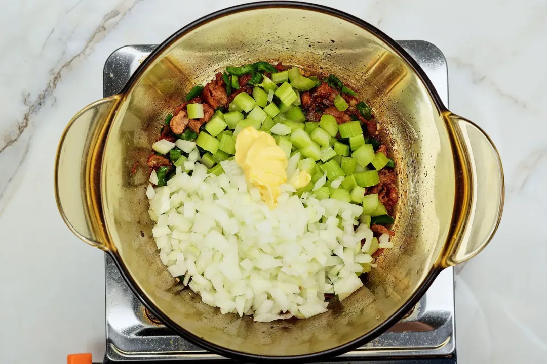 A picture of diced onions, diced celery, minced garlic, cubed jalapeno, butter, and spices being added to a pot of caramelized bacon