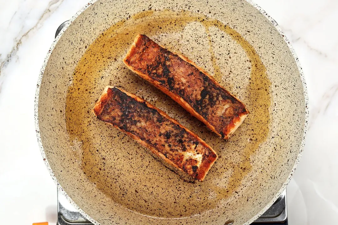 A large pan cooking two salmon fillets whose skin is browned with black charred marks