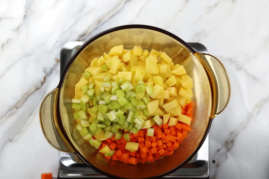 A glass saucepan cooking coarsely-diced potatoes, coarsely-diced carrots, and coarsely-chopped celery
