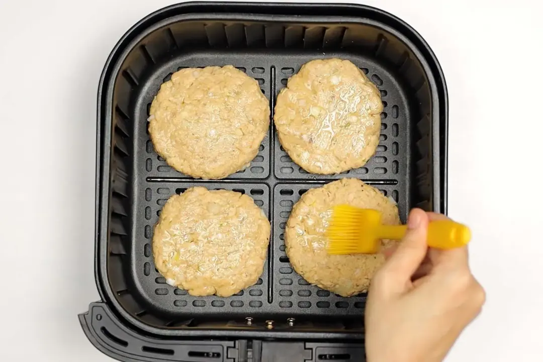 An air fryer basket containing four uncooked burger patties and a hand spreading butter with a yellow butter brush on them