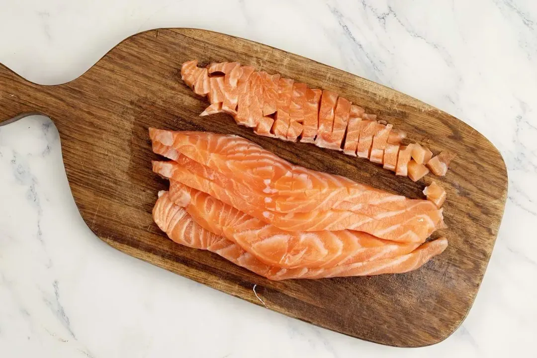 A piece of cured salmon fillet being diced on a wooden cutting board