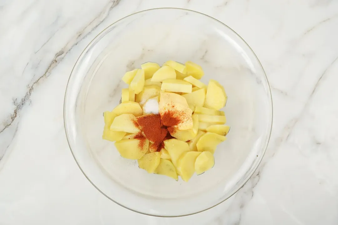 A large glass bowl containing uncooked potato cubes, paprika, salt, and butter