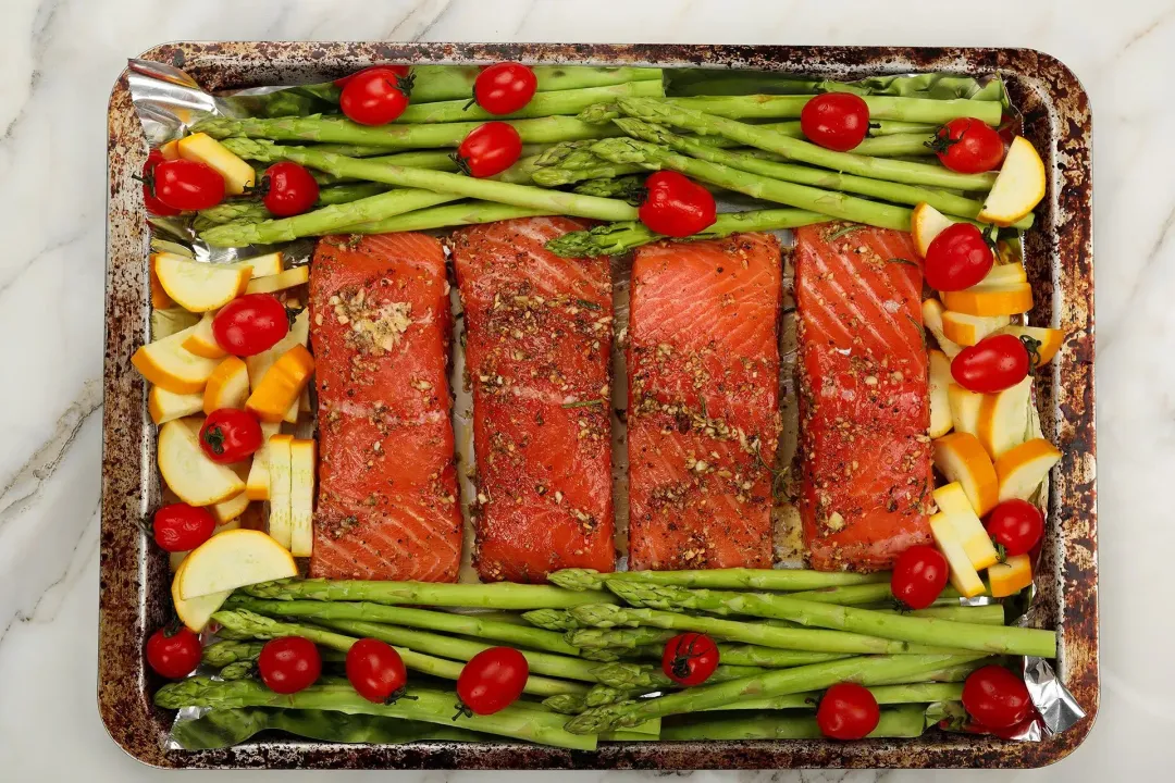 Salmon fillets, asparagus, cherry tomatoes, and slices of zucchini on a baking tray, about to be baked