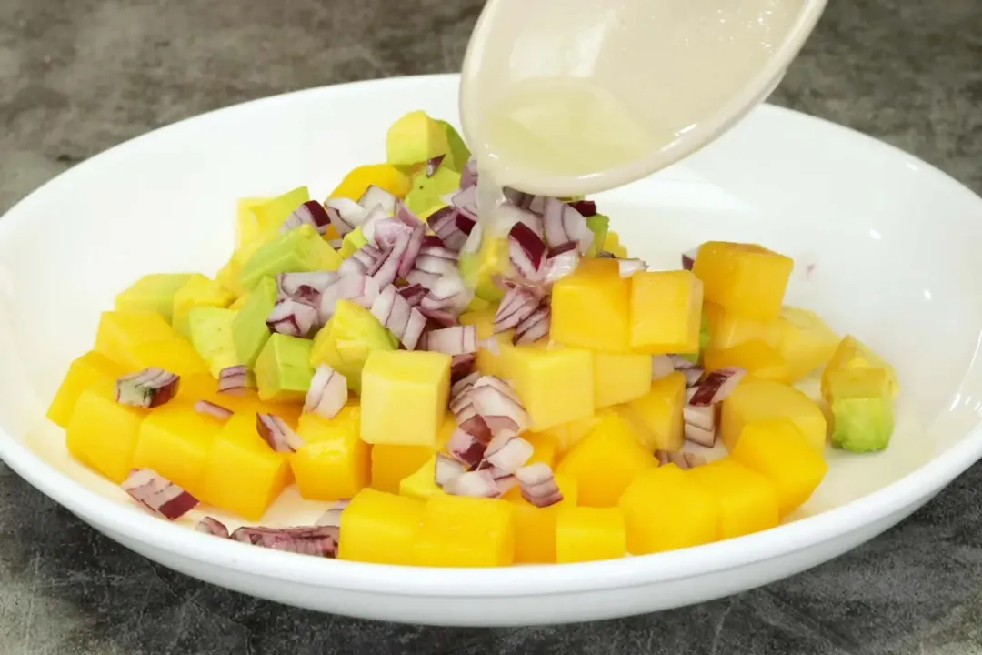 A picture of a spoon drizzling some lime juice over the mixture of mango cubes, avocado cubes, and diced onion.