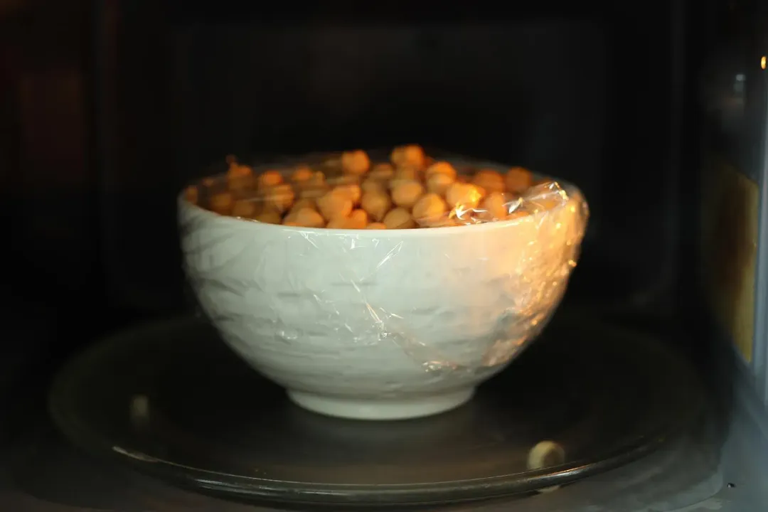A picture of a bowl of sanned chickpeas wrapped in plastic wrap and being microwaved