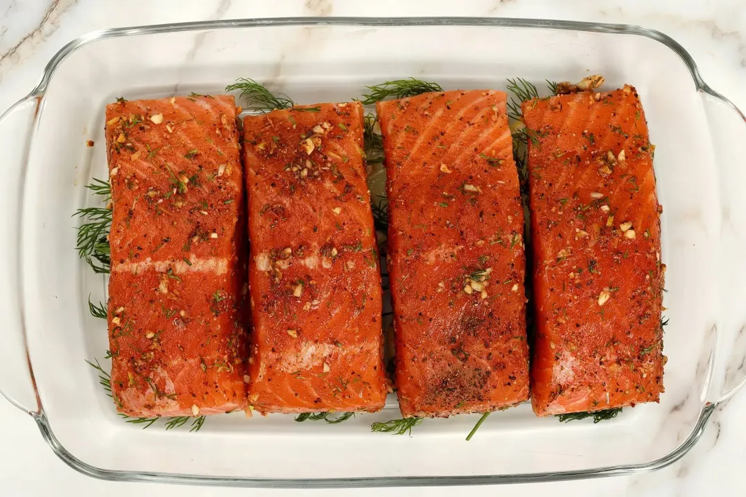 A rectangle glass tray containing four uncooked salmon fillets covered in spices and herbs, laid on a bed fresh dill