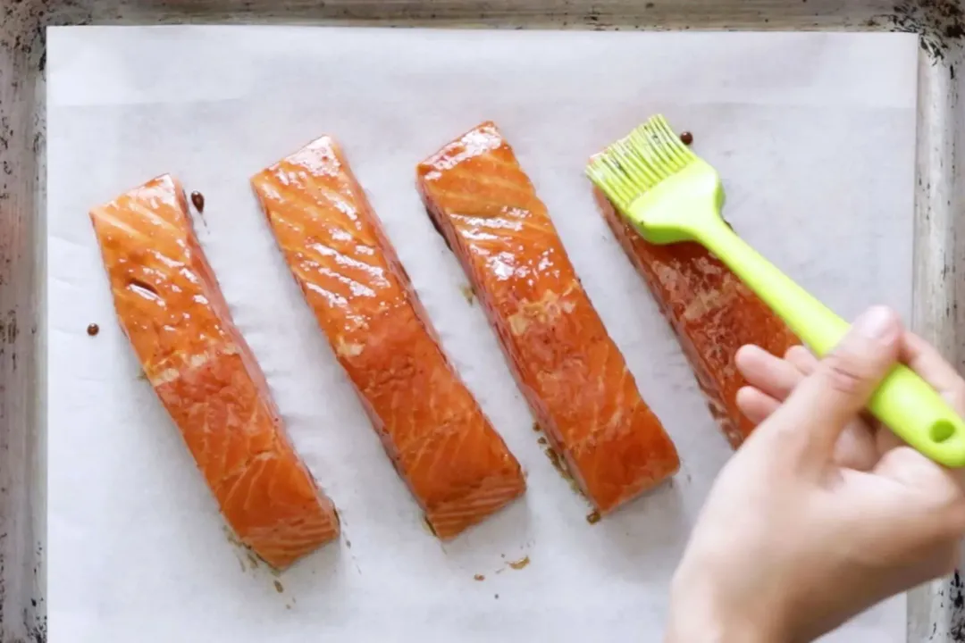 A hand spreading sauce onto four uncooked salmon fillets laid on a baking sheet lined with parchment paper