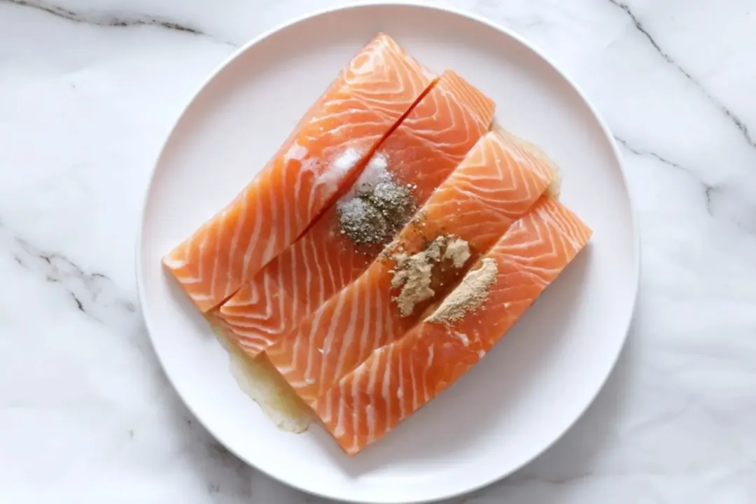 A plate containing four raw salmon fillets with some spices on top