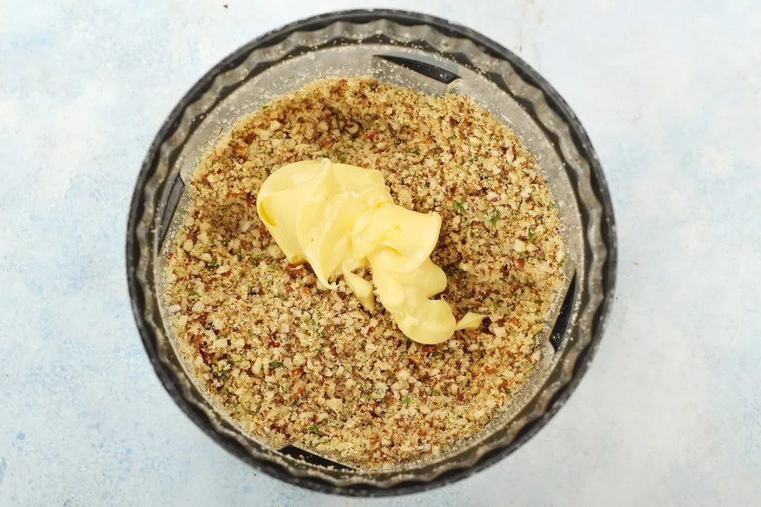 A tablespoon of butter is added to a pecan crust mixture in a food processor.