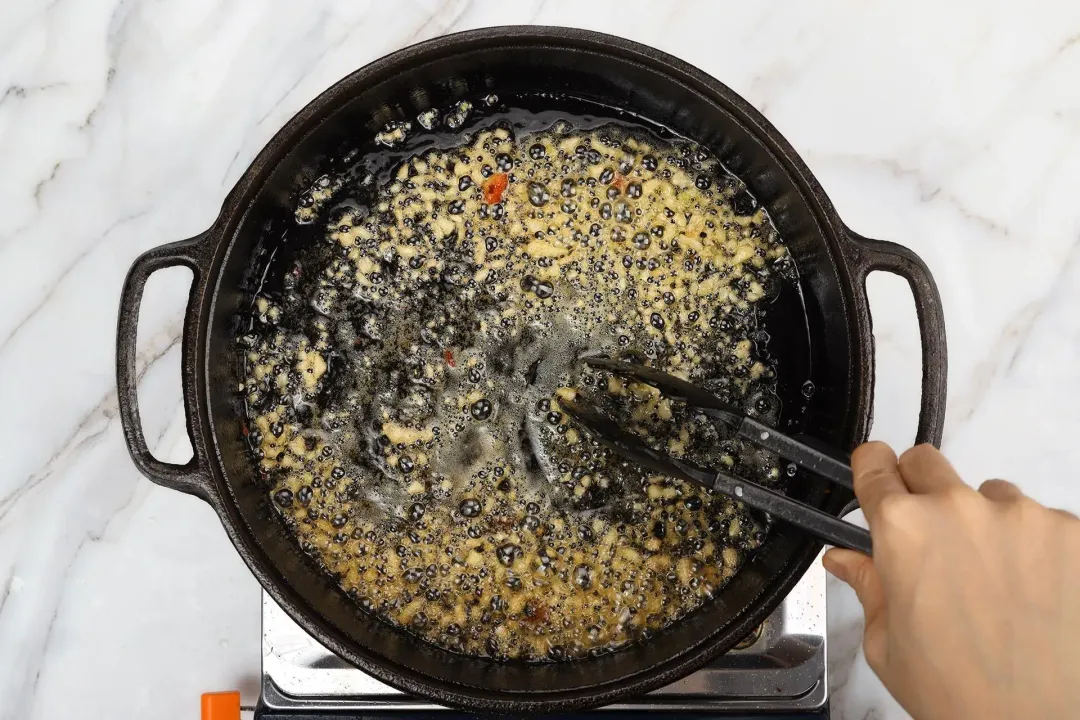 A hand using a pair of tongs to cook finely minced garlic with melted butter in a skillet