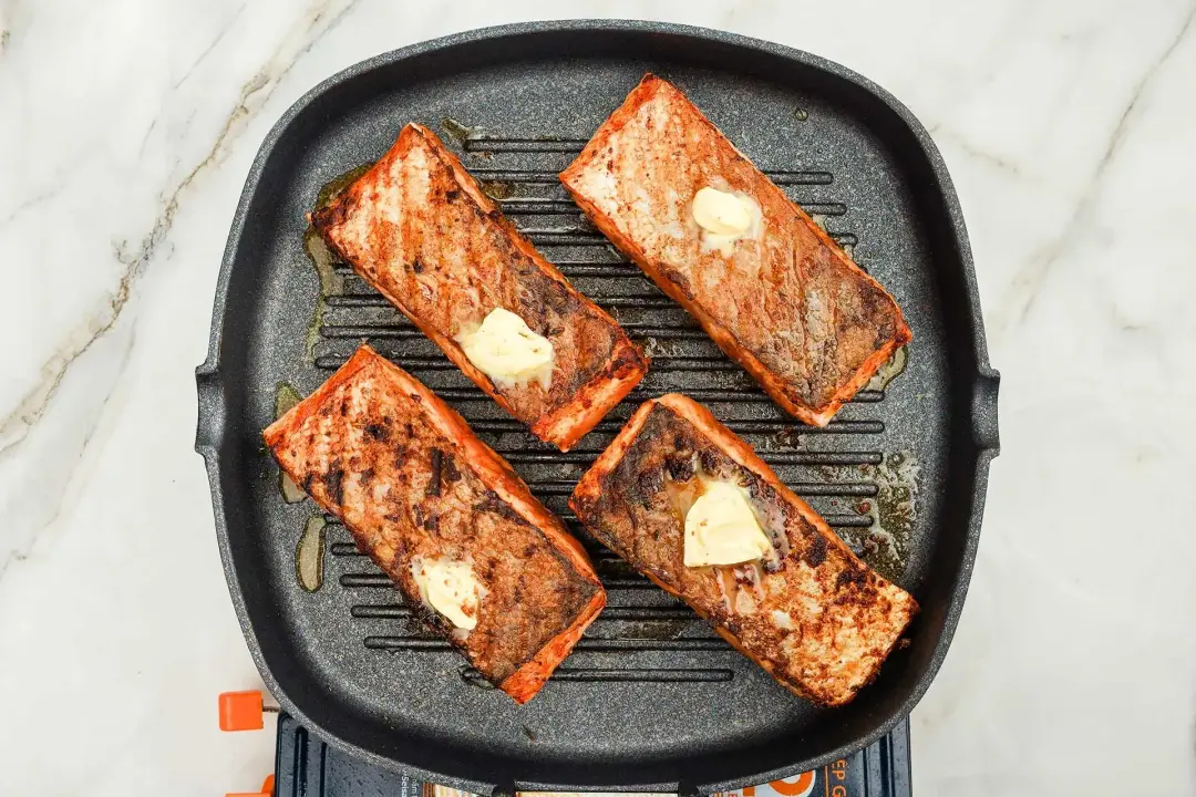 Searing four salmon fillets in a cast iron skillet with butter on top