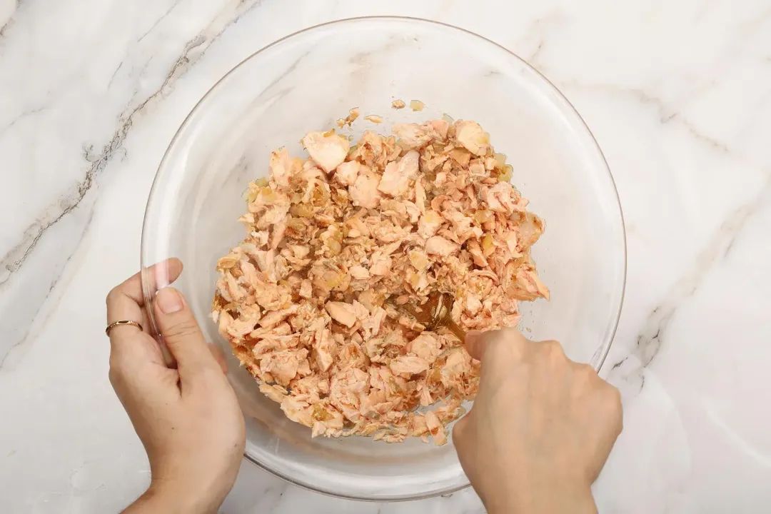 Two hands flaking pink cooked salmon in a large glass bowl with a fork