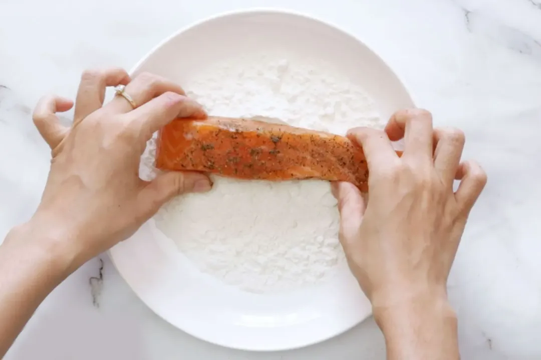 Two hands dipping a salmon filet into a flour plate