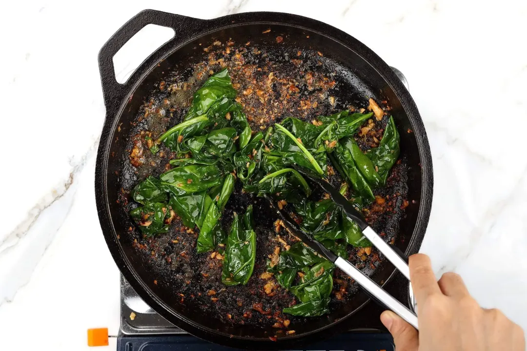 A hand using a pair of tongs to cook spinach leaves on a skillet