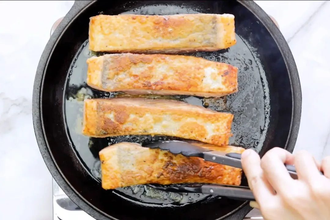 Four salmon filets are being seared with oil in a skillet, one of which is being held by a food grabber.
