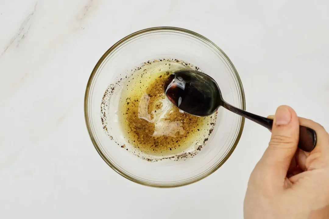 A hand holding a black spoon and stirring a small glass bowl containing a dark yellow dressing that’s mixed with ground black pepper 