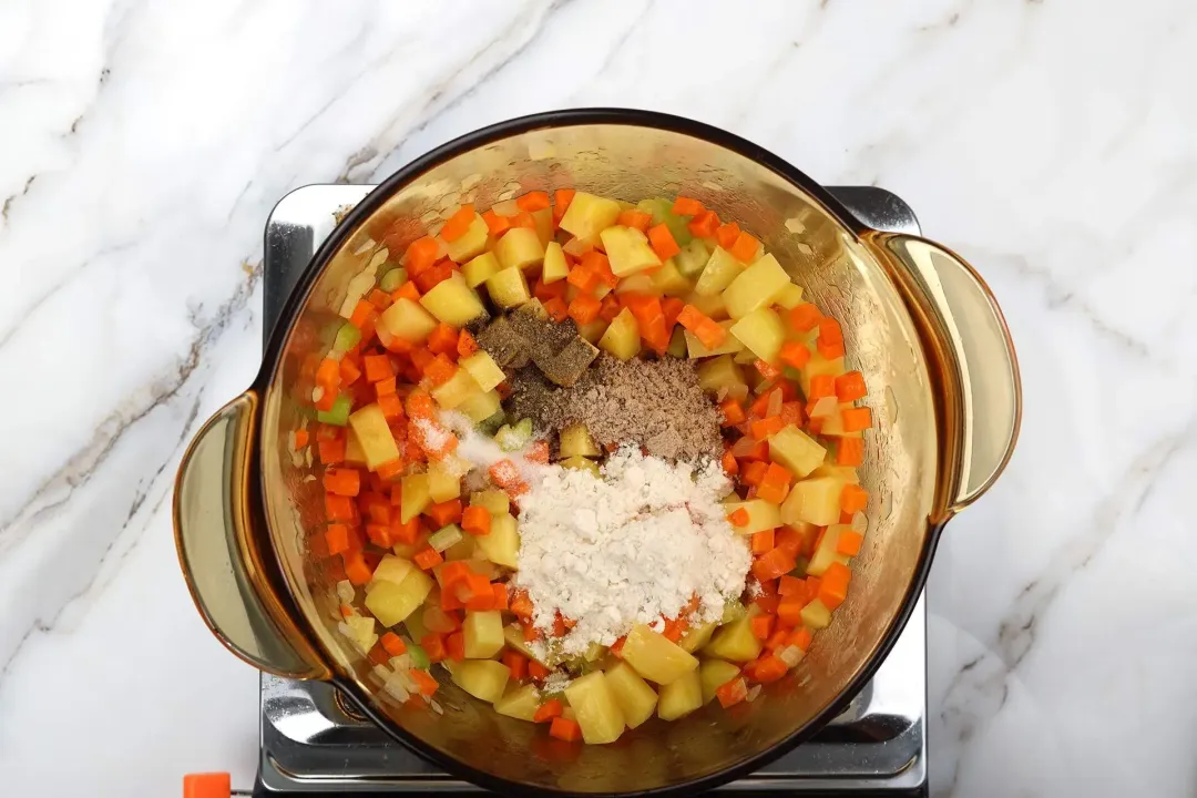 A glass saucepan cooking coarsely-diced carrots and potatoes with some flour, gravy cubes, and black pepper on top