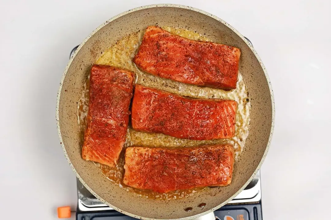 A skillet on a stove with four salmon fillets and some oil in it.