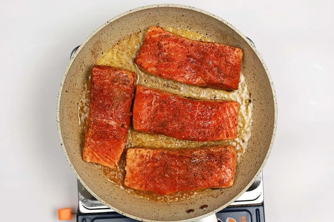 A skillet on a stove with four salmon fillets and some oil in it.