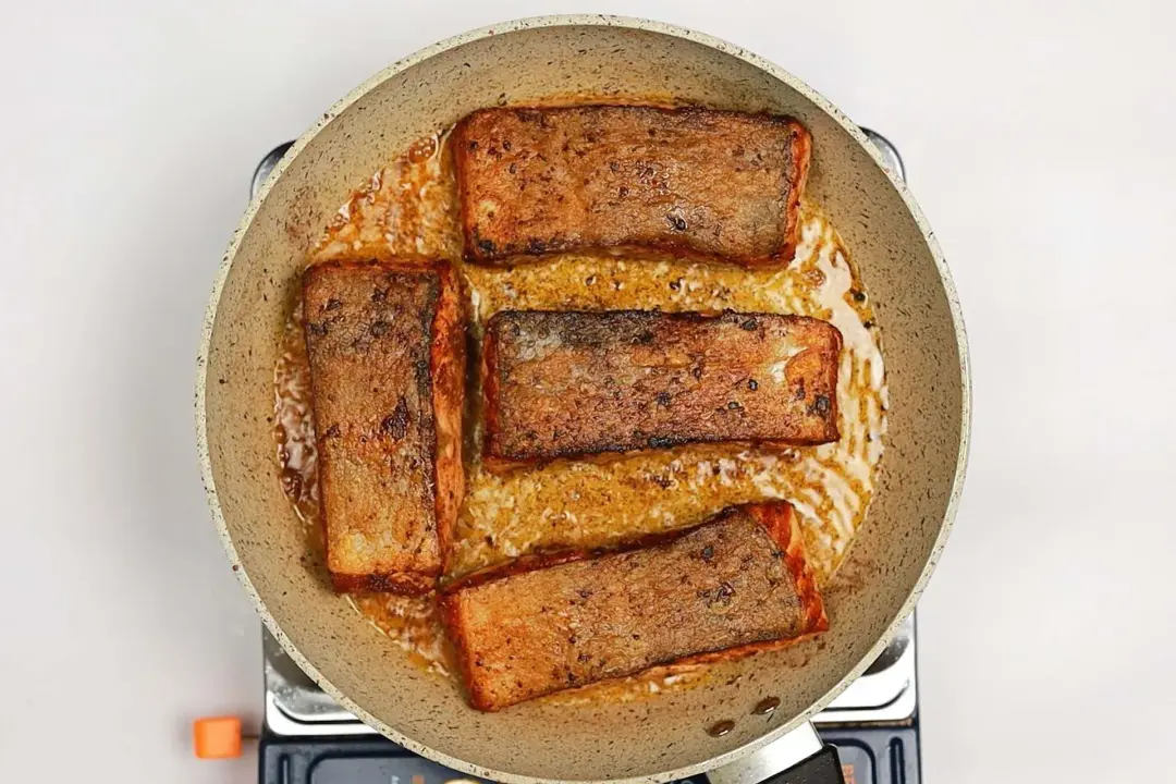 A skillet on a stove with some oil and four salmon fillets skin-side up in it.