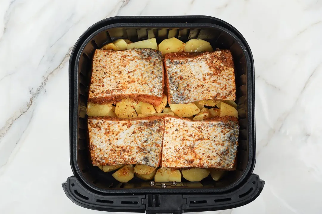 An air fryer basket containing four uncooked salmon fillets laid on a bad of potato cubes