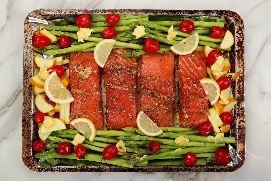 A baking tray of salmon fillets and vegetables with some slices of lemon being scattered on top