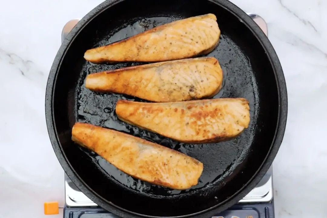 Four salmon filets sizzling in an oiled cast iron skillet