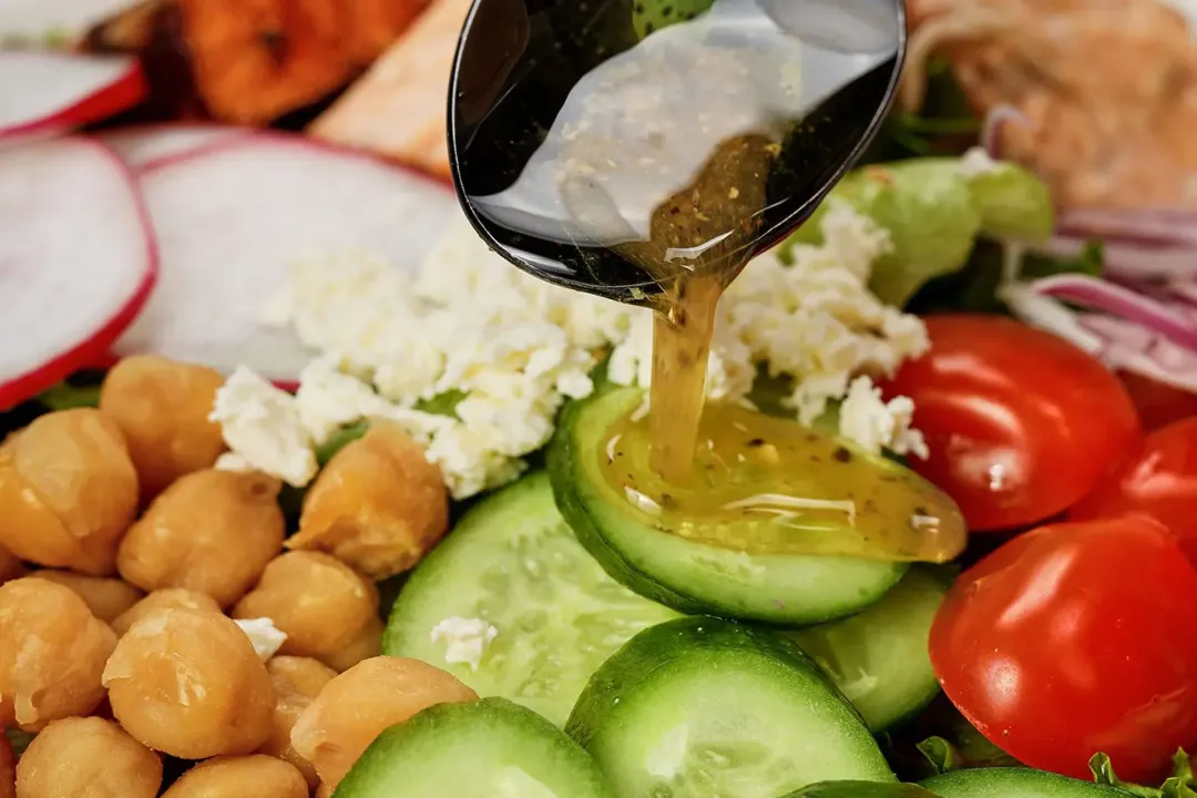 A black spoon dispensing a syrupy and yellow dressing onto slices of cucumbers, chickpeas, feta cheese, and cherry tomatoes of a salad