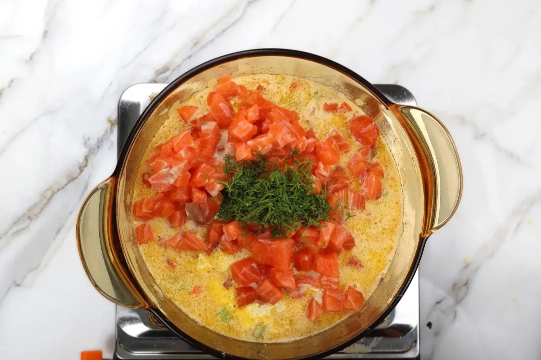 A glass saucepan cooking a soup with bright orange uncooked salmon cubes and chopped dill