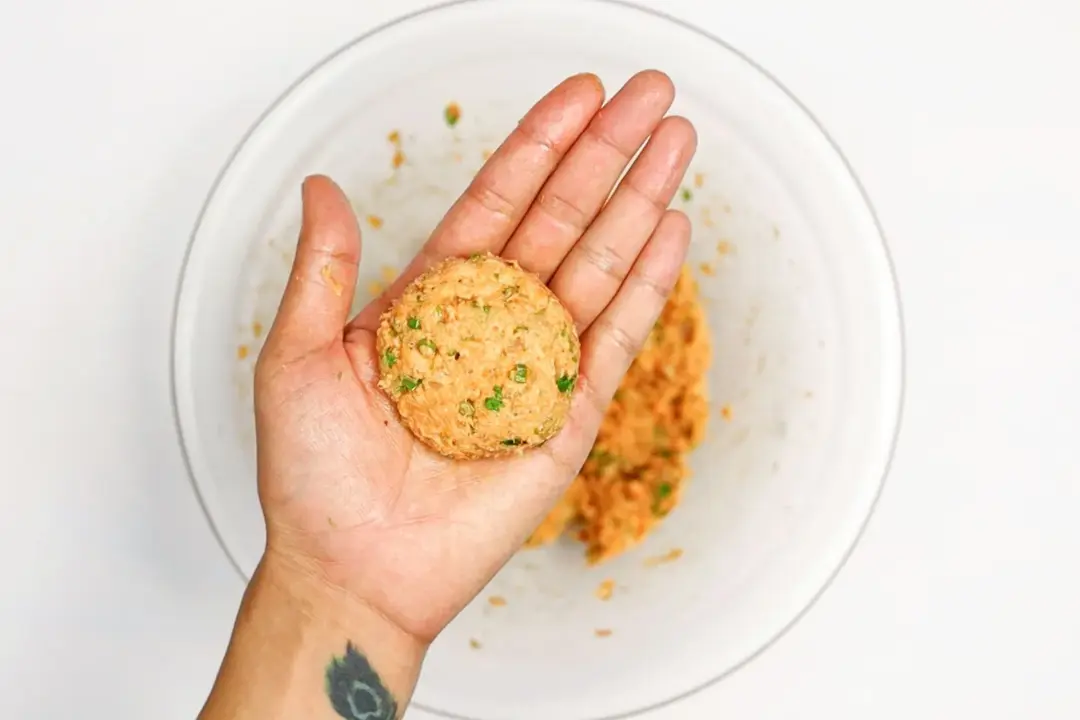 An uncooked salmon patty in the palm of a hand in the background of a glass bowl of salmon patties mixture.