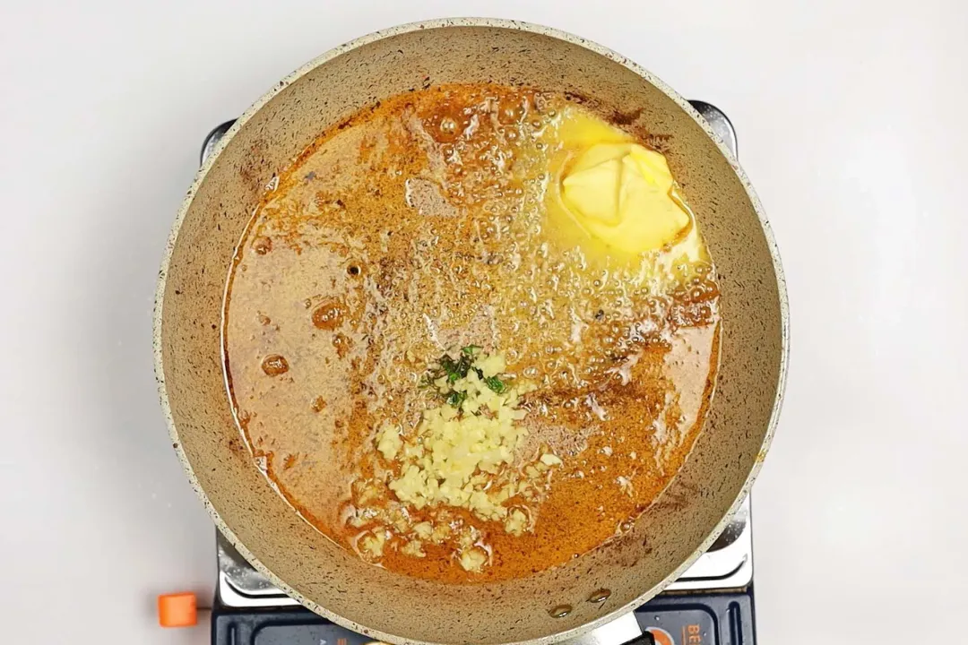 A skillet on a stove with some bubbling oil, thyme, butter, and minced garlic in it.