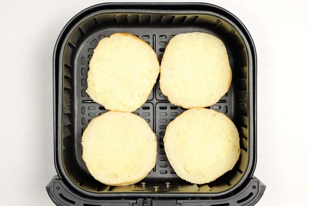 An air fryer basket containing four hamburger buns with the cut side facing upward