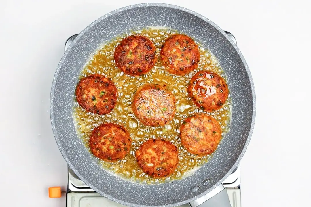 A skillet on the stove with bubbling oil and eight salmon patties in it.