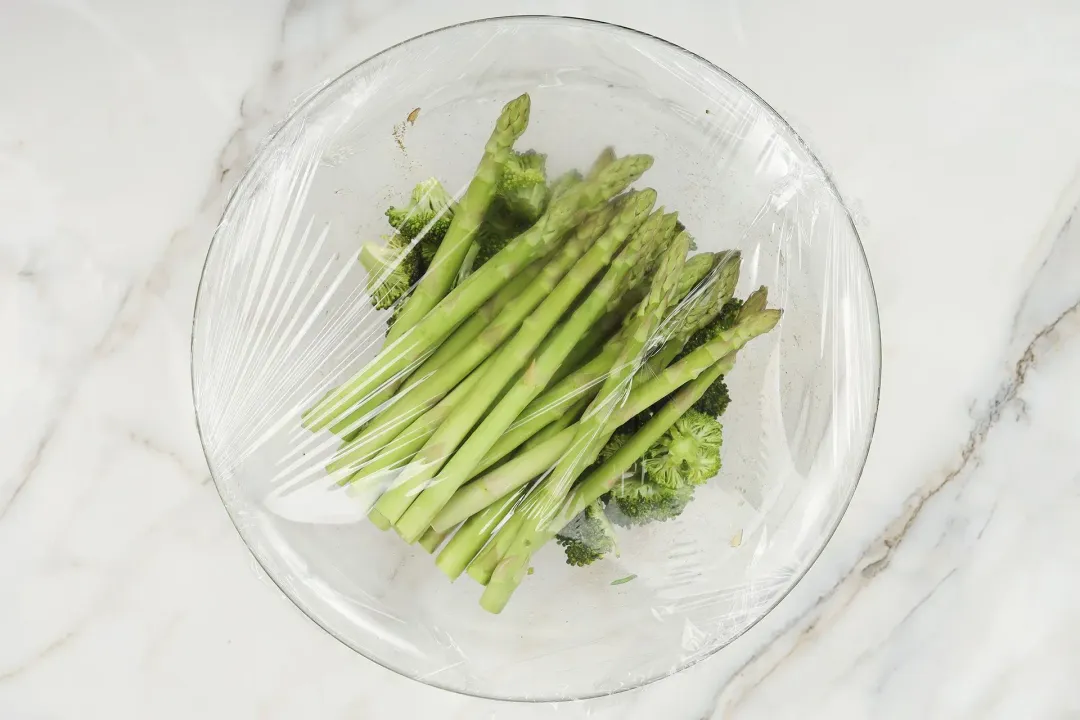A large glass bowl containing asparagus and broccoli florets covered in cling wrap