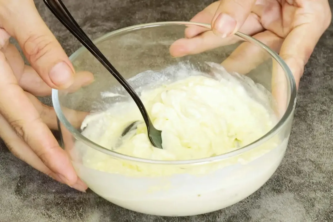 Two hands holding a glass bowl of a white sauce with a spoon in it.