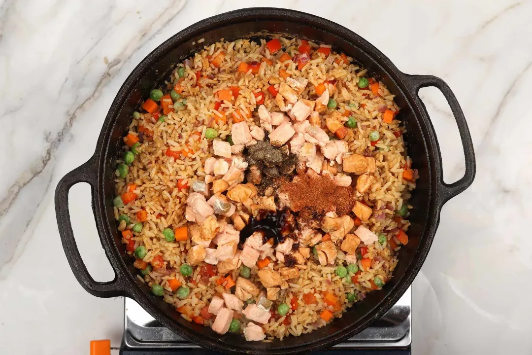 Cubed salmon and seasonings added to a colorful veggie fried rice in a black cast iron skillet