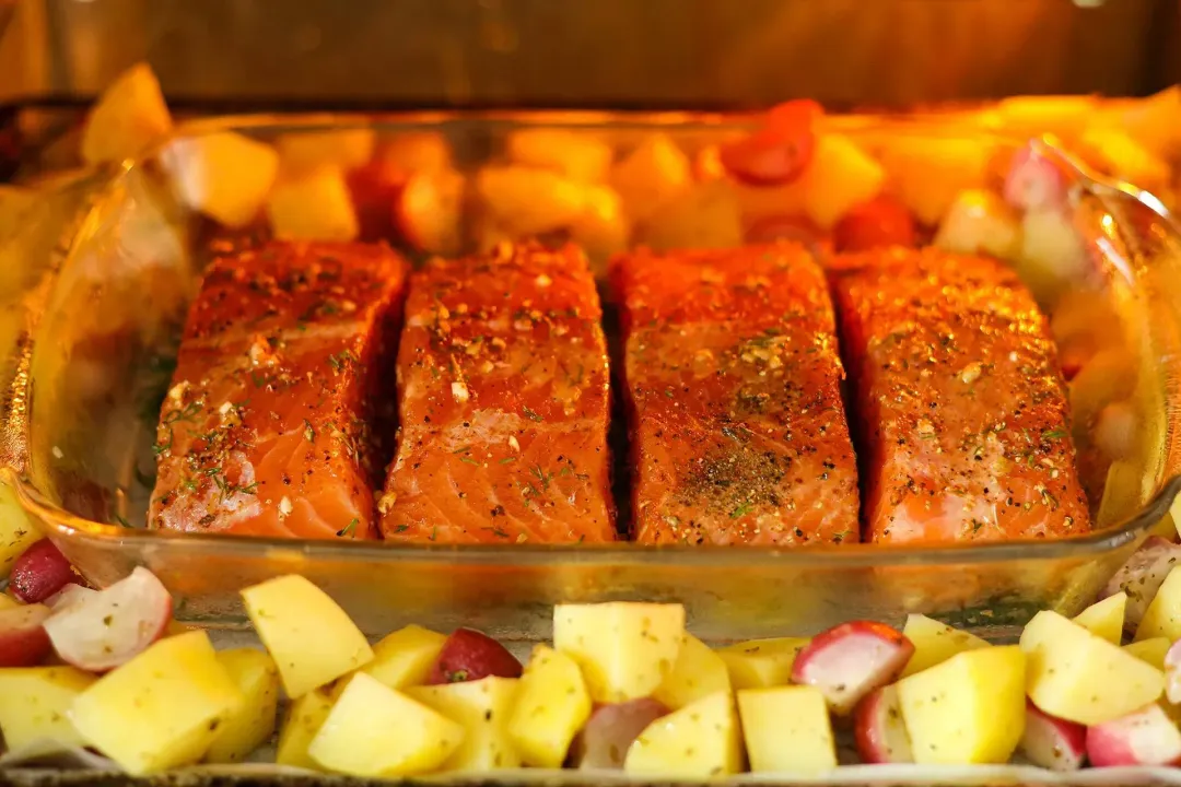A glass tray containing four salmon fillets surrounded by potato cubes and radish cubes being cooked in an oven