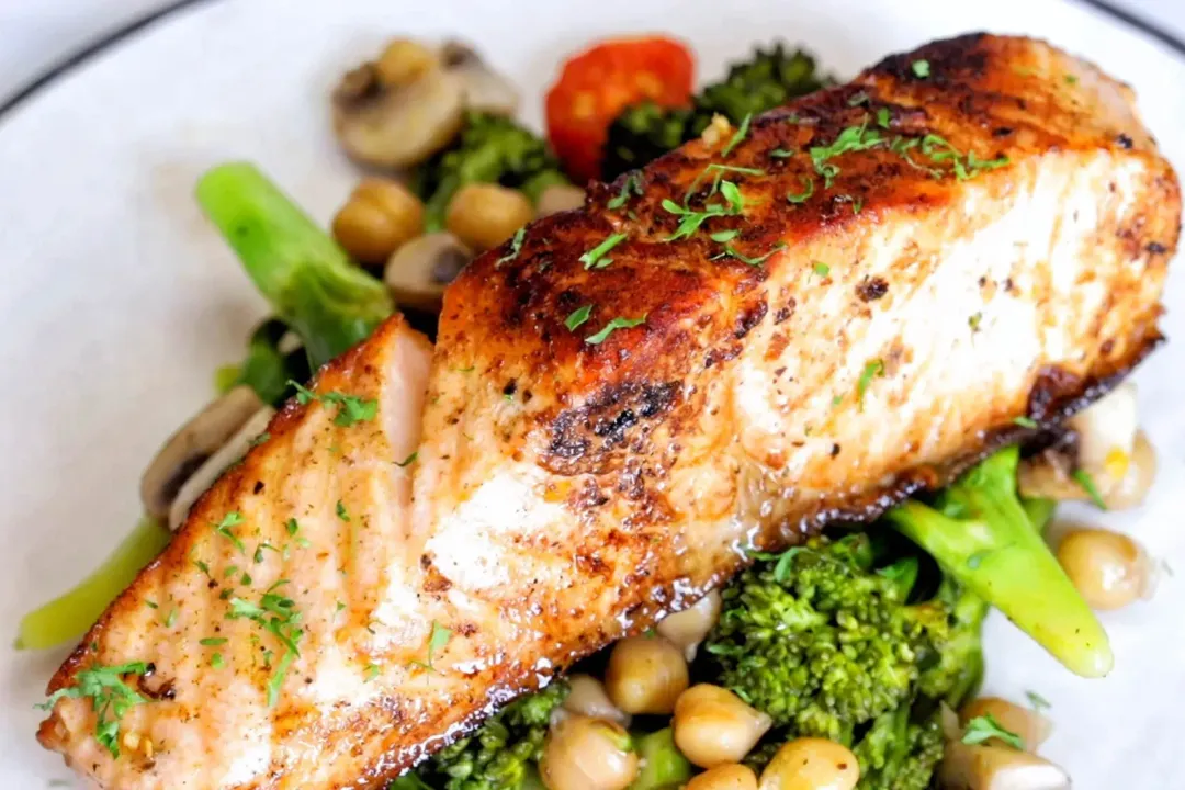 A close-up shot of a pan-seared salmon fillet laid on a bed of broccolini and chickpeas, topped with some chopped parsley