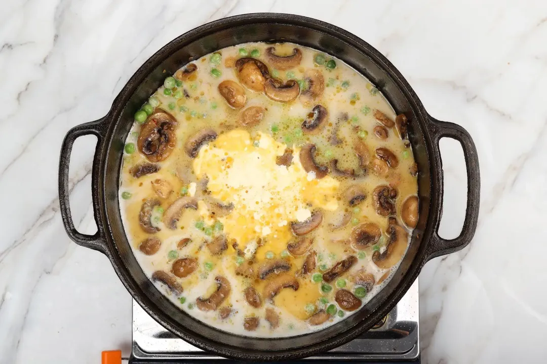 A skillet cooking browned mushrooms and green peas in heavy cream with yellow mustard