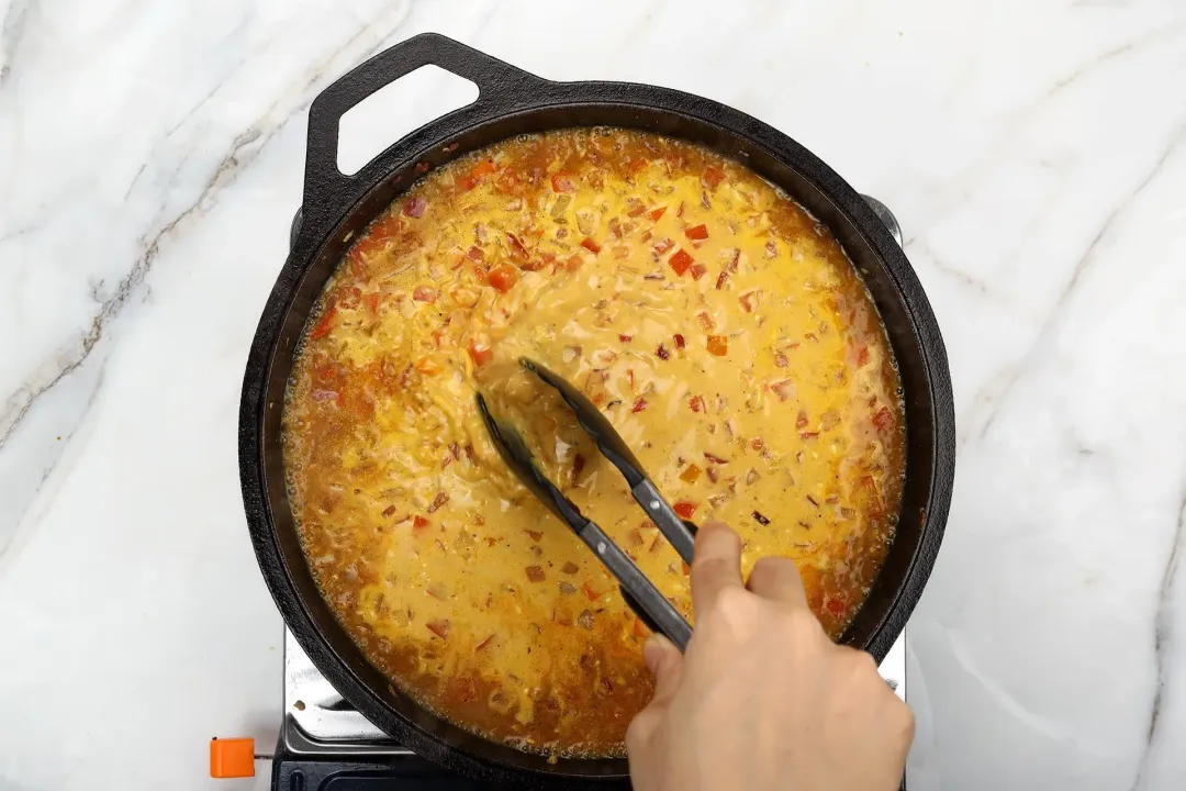 A hand holding a pair of tongs stirring a skillet filled with a dark yellow curry