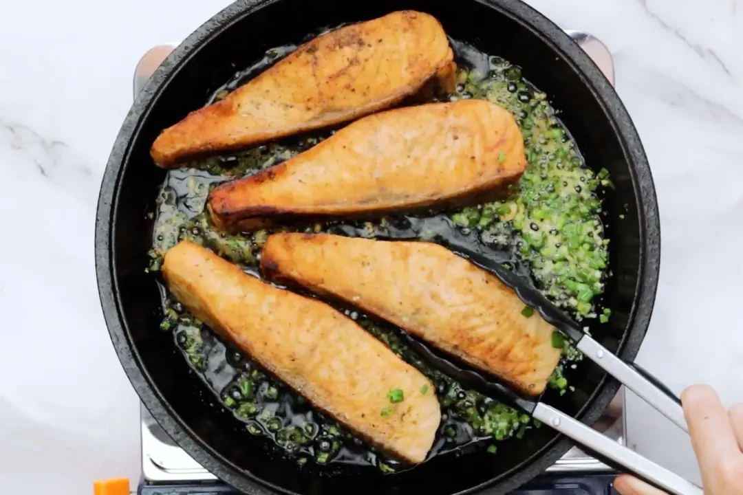 A black cast iron skillet with hot oil underneath sizzling salmon filets and some chopped herbs