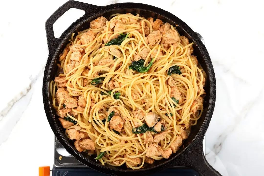 A cast iron skillet filled with cooked spaghetti, salmon, and spinach laid near a striped tablecloth