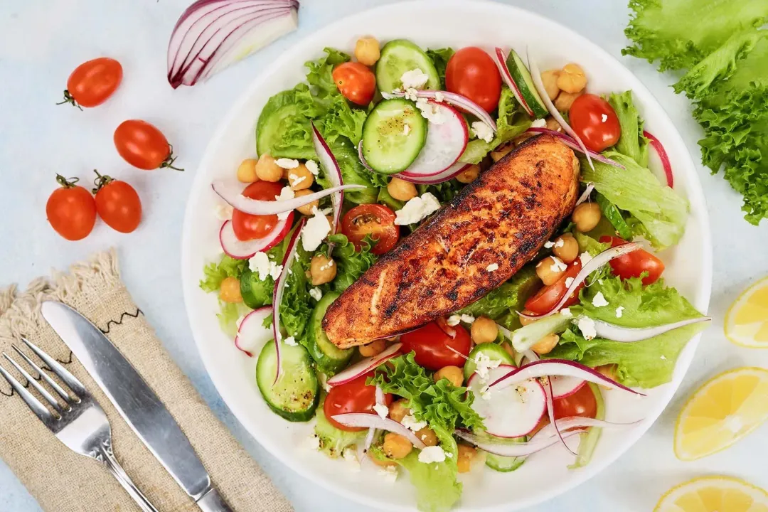 A round white plate containing a salmon salad laid near lemon wedges, cherry tomatoes, lettuce leaves, a beige tablecloth, a dinner knife, and a fork